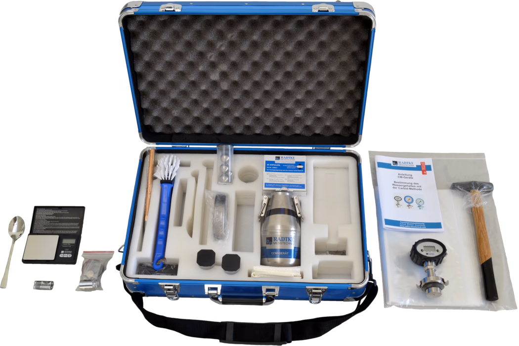 Offener blauer Transportkoffer mit Schaumstoffeinlage, der ein Feuchtemess-Kit enthält. Sichtbar sind ein Metall-Probenbehälter mit Aufschrift 'RADTKE MESSTECHNIK', eine Langbürste, eine kleine digitale Waage mit Löffel, eine Bedienungsanleitung, ein Druckmanometer, ein Holzhammer, Batterien, verschiedene versiegelte Kleinteile und mehrere schwarze Schutzkappen, alles sauber in ausgeschnittenen Schaumstofffächern angeordnet.