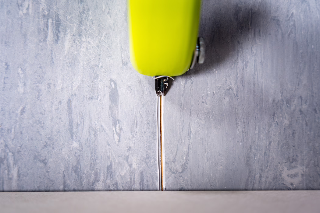 WOLFF_Application Image_Groover_2024-01 (12) Nahaufnahme eines limonengrünen, einziehbaren Cuttermessers, das vertikal über einer schmalen Fuge zwischen einer strukturierten grauen Wand und einem hellen Boden positioniert ist. Die dünne Metallklinge ist nach unten in die Fuge ausgefahren, der Kunststoffgriff und eine kleine Metallschraube sind oben im Bild sichtbar. Die Aufnahme betont kontrastierende Oberflächen — raue, verputzte Wand und glatter Boden — und ist weich von oben beleuchtet, wodurch feine Kratzer und Gebrauchsspuren an Klinge und Griff sichtbar werden.