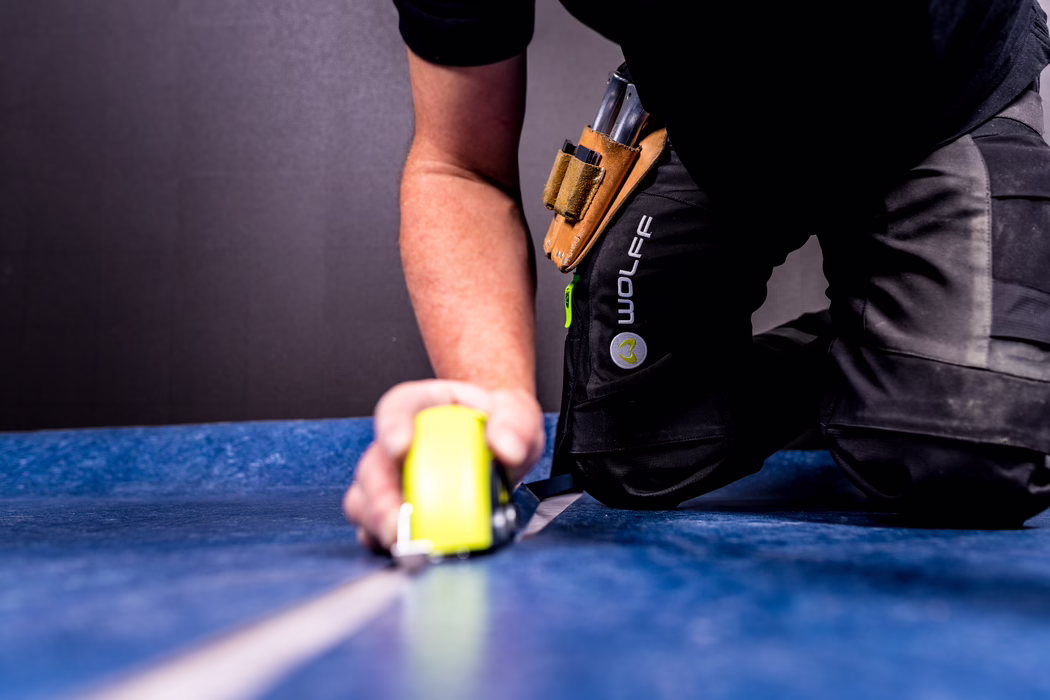 Nahaufnahme eines Arbeiters, der auf einem blauen Boden kniet und mit einem gelben Maßband misst; sichtbare Arbeitshose mit Werkzeugtasche und 'WOLFFE'-Logo, Hand hält das Maßband, graue Wand im Hintergrund.