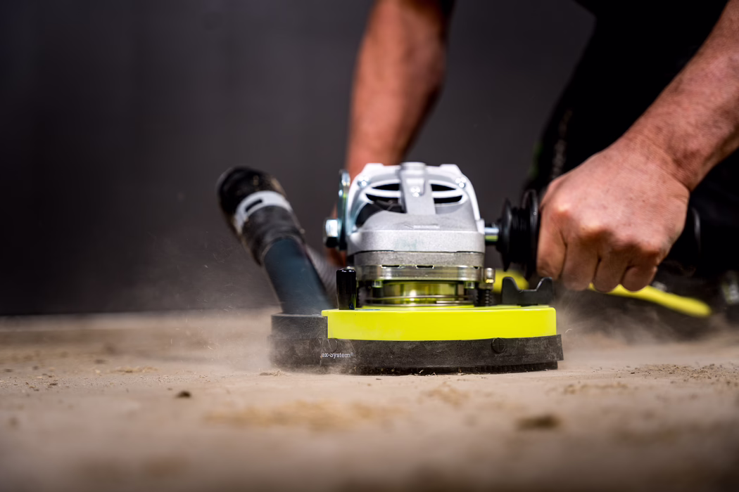 WOLFF_Application Images_BS 180_2024-01 (18) Nahaufnahme eines Handwerkers, der eine handgeführte Bodenschleifmaschine auf einer Betonoberfläche bedient; der gelbe Schleifkopf dreht sich und wirbelt Staub auf, während kräftige Hände die Griffe halten.
