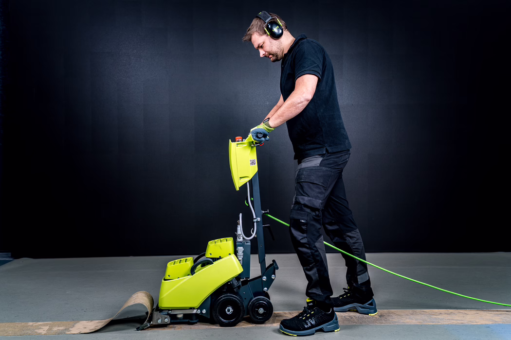 WOLFF_Application Image_VarioSilent_Detail1_2022-11 Ein Mann mit Gehörschutz und Handschuhen bedient eine gelbgrüne Bodenschleifmaschine auf einem grauen Boden und rollt einen Streifen alten Bodenbelags auf; er trägt schwarze Arbeitskleidung und Sicherheitsschuhe und konzentriert sich auf die Führung der kompakten elektrischen Maschine, die über ein grünes Stromkabel verbunden ist.