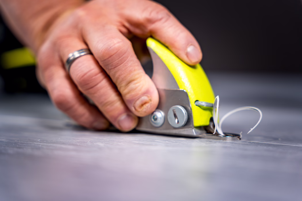WOLFF_Application Image_Groover_2024-01 (1) Close-up of an adult hand using a small handheld cutting tool on a flat gray surface. The bright yellow and metal tool is pressed down by the fingertips, and a thin white curled shaving of material is visible where the blade meets the surface. The hand shows textured skin, short nails and a silver wedding band on the ring finger. Shallow depth of field keeps the focus on the tool and the fingertips while the foreground and background are softly blurred; lighting is soft and even, emphasizing the contrast between the yellow tool, skin tones and the neutral work surface.