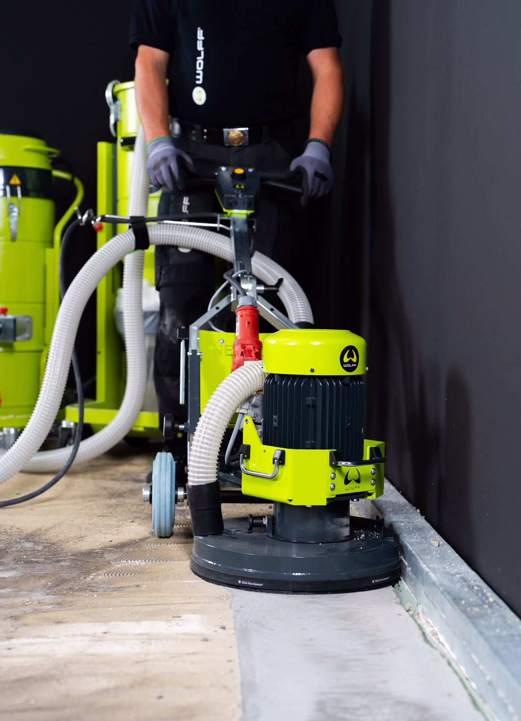 Ein Arbeiter bedient einen leuchtend limonengrünen Industrietrockenschleifer neben einer dunklen Wand. Die Nahaufnahme zeigt den rotierenden Schleifkopf der Maschine, große Absaugschläuche, die an eine Staubabsaugung angeschlossen sind, Gummiräder und ein lenkbares Gestänge, das der Bediener mit Handschuhen hält. Der Holzboden weist Schleifspuren auf und zeigt eine geglättete Betonfläche, wo die Oberfläche entfernt wurde. Der Arbeiter trägt ein schwarzes Shirt und Schutzhandschuhe; das Gesicht ist nicht sichtbar. Die Szene wirkt wie eine Innenrenovierung oder Baustelle.