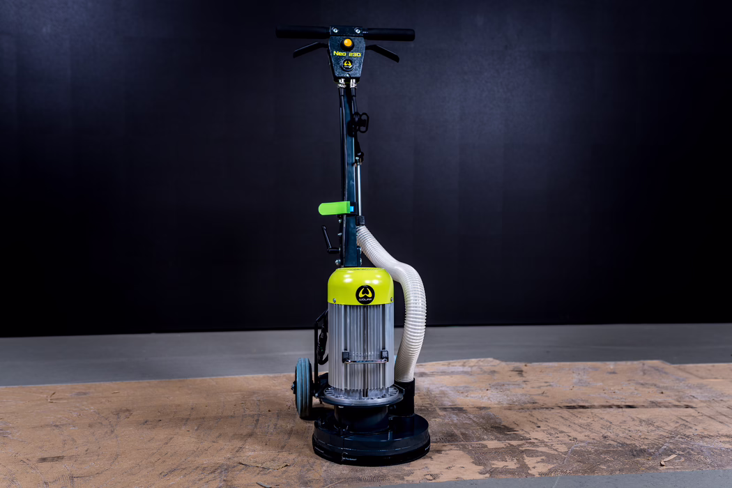WOLFF_Application Image_Neo230_Main_2022-11 Eine einmotorige Scheuerscheiben-/Schleifmaschine, frontal und aufrecht in einem Studio mit dunklem Hintergrund fotografiert; im Vordergrund liegen abgenutzte Holzbohlen. Die Maschine besitzt ein zylindrisches Motorgehäuse mit auffälliger gelb-grüner Abdeckung, einen vertikalen Lenker mit horizontalem Griff, zwei kleine Transportrollen hinten und einen flexiblen weißen Staubschlauch, der vom Motor zur Schleifplatte führt. Die runde Schleifbasis steht auf dem Holz, Studiobeleuchtung betont Metalloberflächen, Schlauch und Bedienelemente, während der Hintergrund gleichmäßig dunkel bleibt.